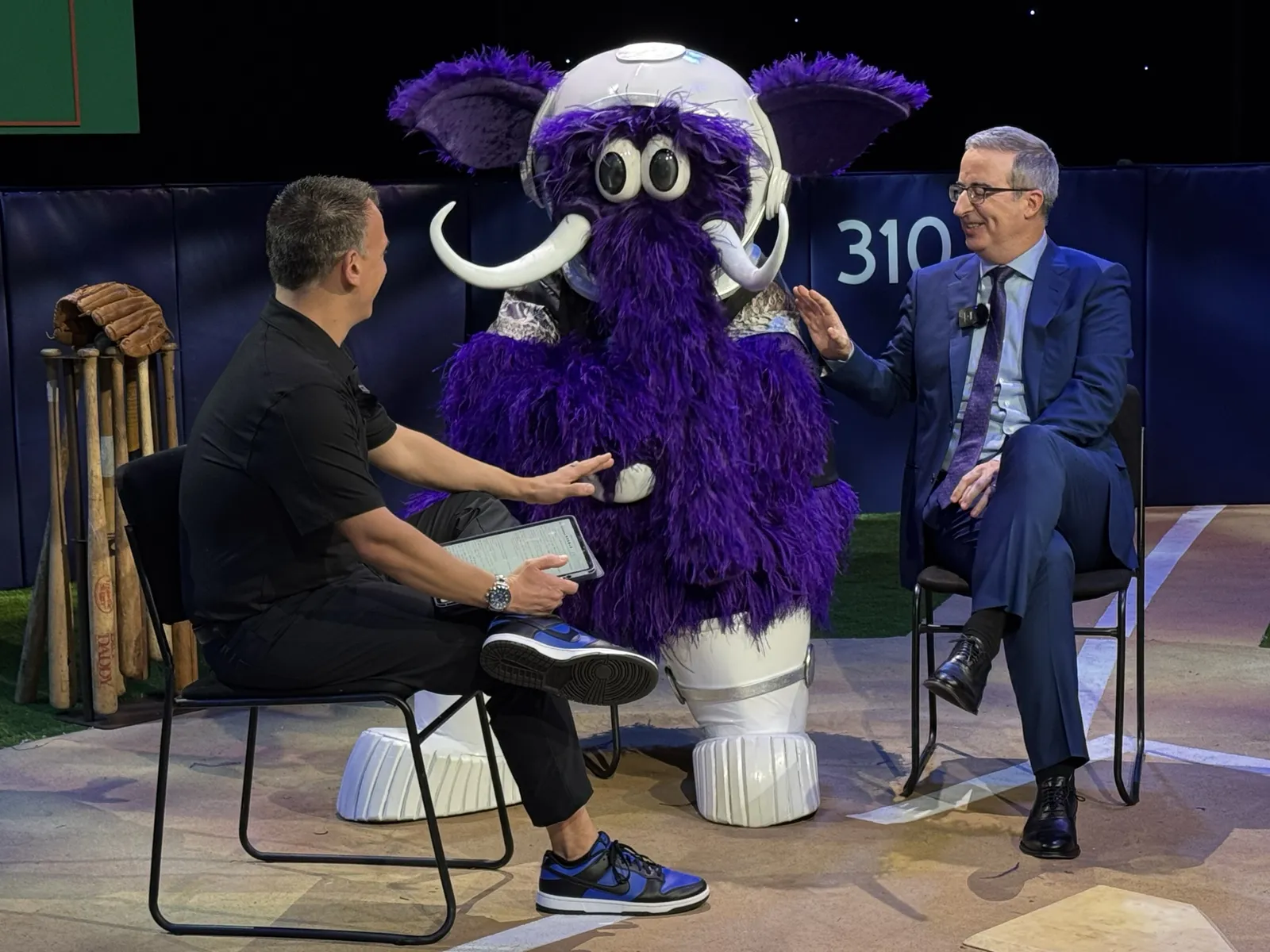 Greg Gania interviewing HBO’s John Oliver on stage beside a mascot
